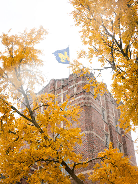 The flag on top of the Michigan Union blows in the wind during the first snowfall of Fall 2025. The bright yellow leaves on a tree nearby sit in stark contrast to the bright white sky.