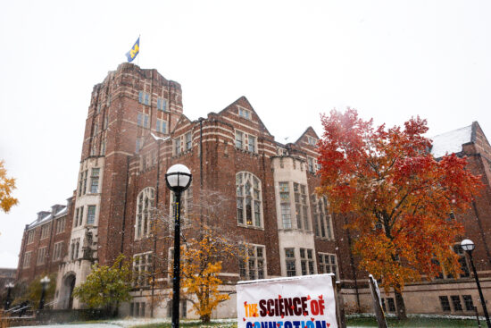 The flag on top of the Michigan Union blows in the wind during the first snowfall of Fall 2025. The rooftops of the building are all dusted in snow, while the trees in the lawn still hold onto their bright red and orange leaves.