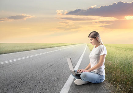 Smiling young woman sitting on a countryside road and using a laptop (stock image)