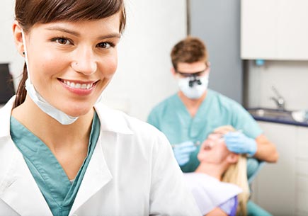 A portrait of a dental assistant smiling at the camera with the dentist working in the background. (stock image)