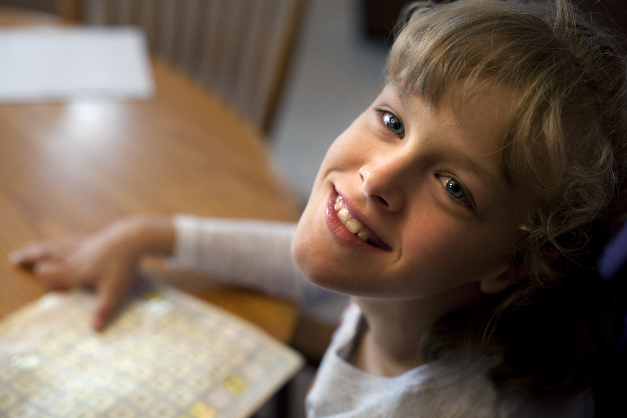 Sitting by the dining table in her parents' Westphalia, MI house, Grace smiles to the photographer. Her hand rests on a board with letters and numbers she uses to communicate. Image credit: Marcin Szczepanski
