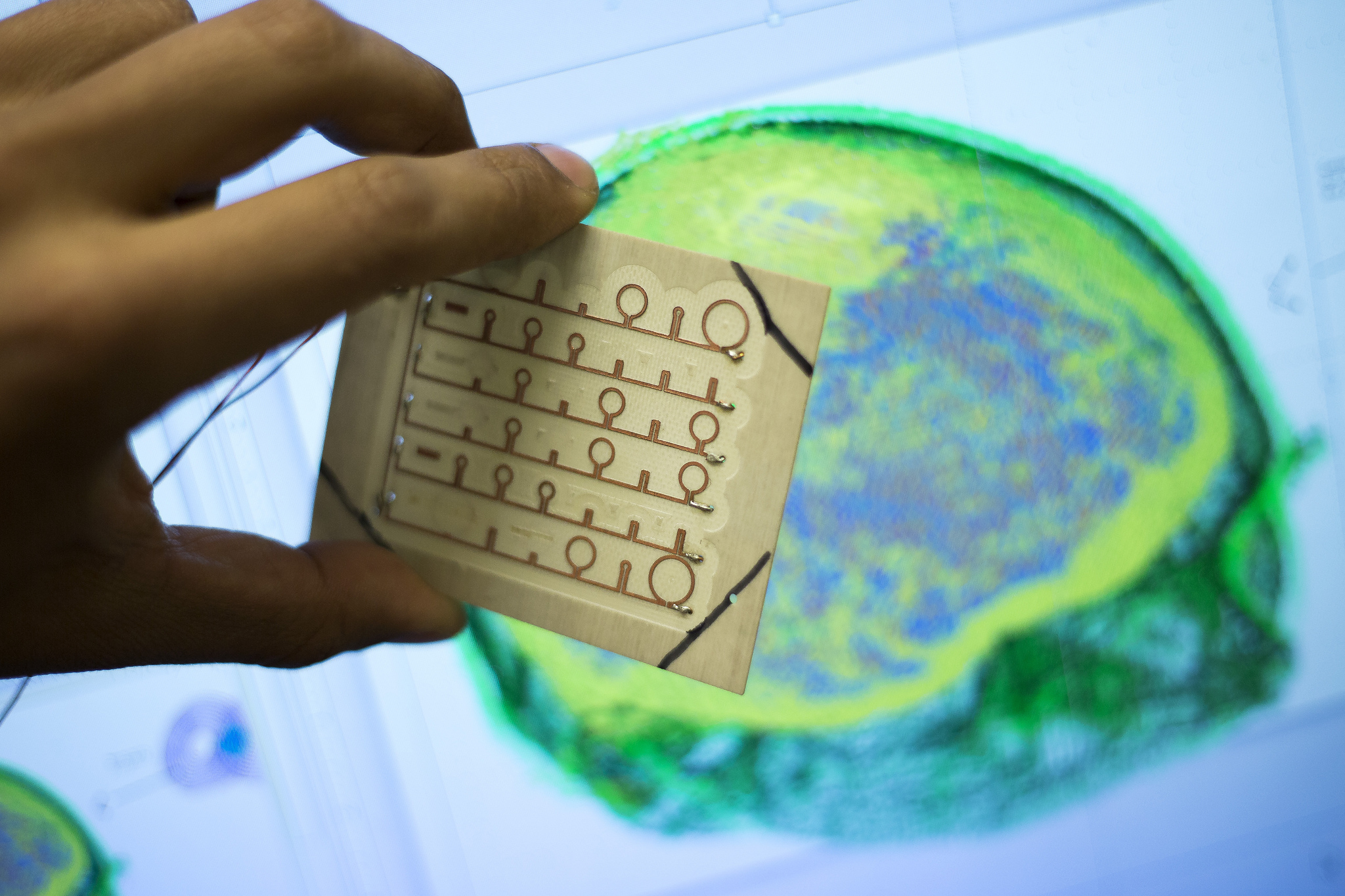 Luis Gomez, Electrical Engineering PhD Student, shows the headpiece -- a square array of 64 circular metallic coils -- next to computer simulations of a brain stimulation in the EECS Building on October 28, 2013.  Researchers and doctors hope that the technology can one day help hit finer targets in the brain that are twice as deep as they can reach today to help improve depression treatment. Image credit: Joseph Xu, Michigan Engineering Communications & Marketing