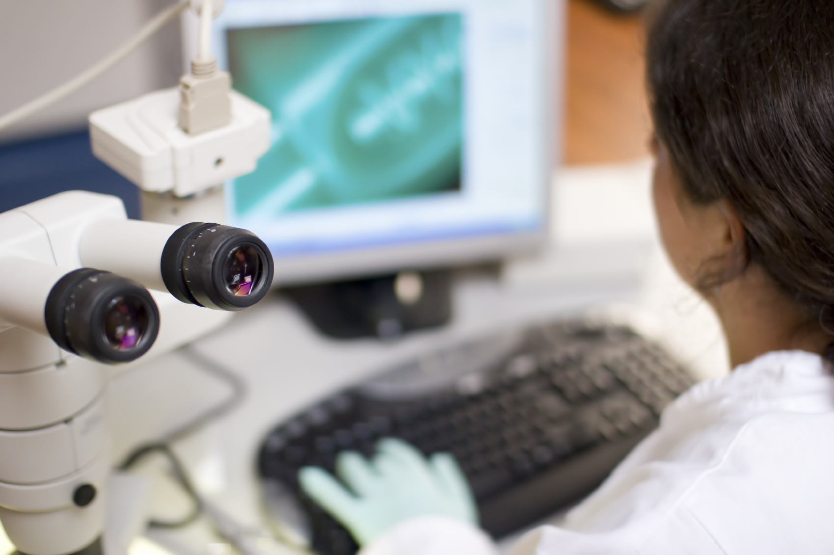 Out of focus view over the shoulder of a researcher using a computer and microscope. (stock image)
