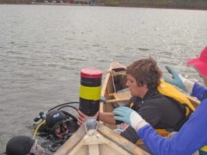 Scuba divers hand a coring device containing lake-bottom sediments to U-M biologist Mary Rogalski at a Connecticut lake. Image credit: Phoebe Zarnetske
