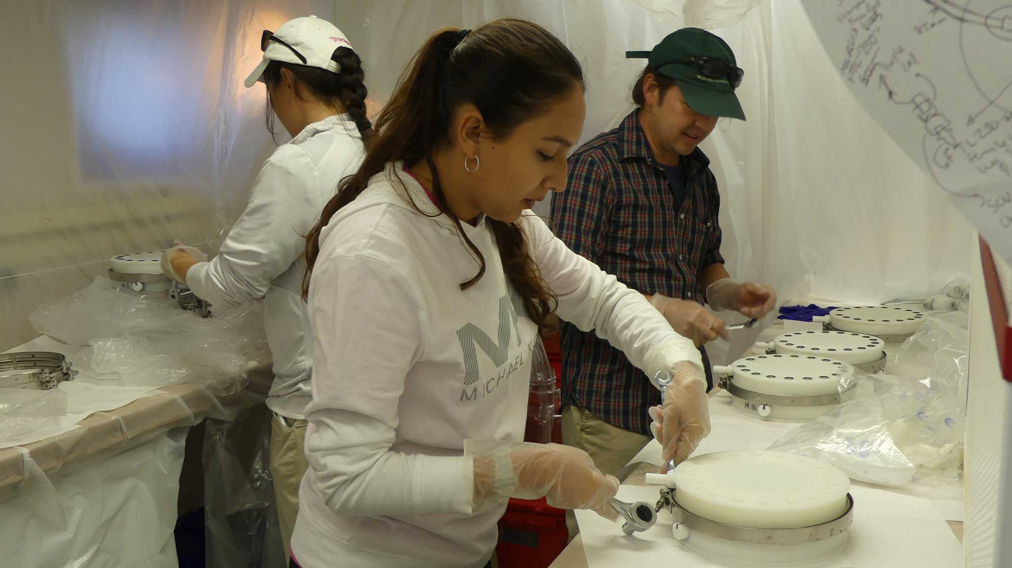 WHOI biogeochemist&nbsp;Mak Saito (far right), Brianna Alanis of UTRGV (center), and Sharon Grim of Michigan open samples collected by Clio to prepare them for microbiological and biochemical analysis inside a "clean lab." To prevent contamination, researchers work within a temporary plastic cleanroom environment lab onboard the ship. Image courtesy: Abigail Noble