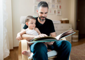 A mid adult father sitting on a chair with baby on lap at home and reading a book in a horizontal medium shot indoors.