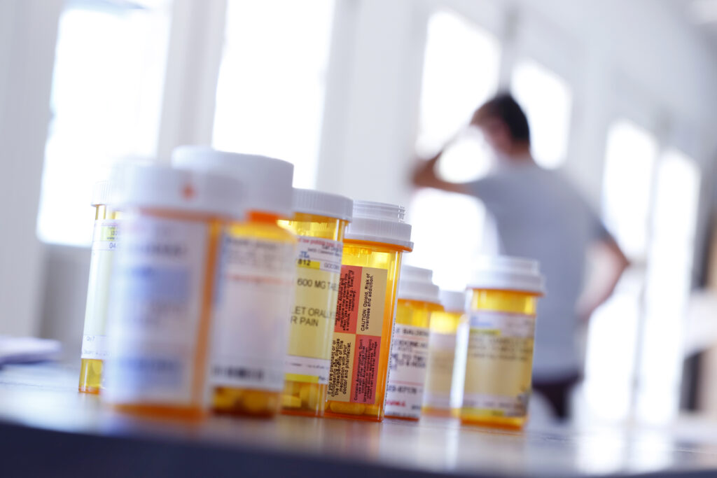A large group of prescription medication bottles sit on a table as a man in the background leans against a wall with his hand on his head.. The image is photographed with a very shallow depth of field with the focus being on the pill bottles in the foreground.