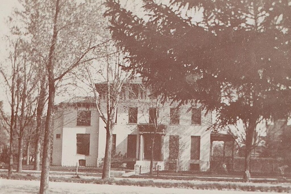 The front of the house in the Angell era, with a low decorative fence, a large evergreen at right and many young deciduous trees.
