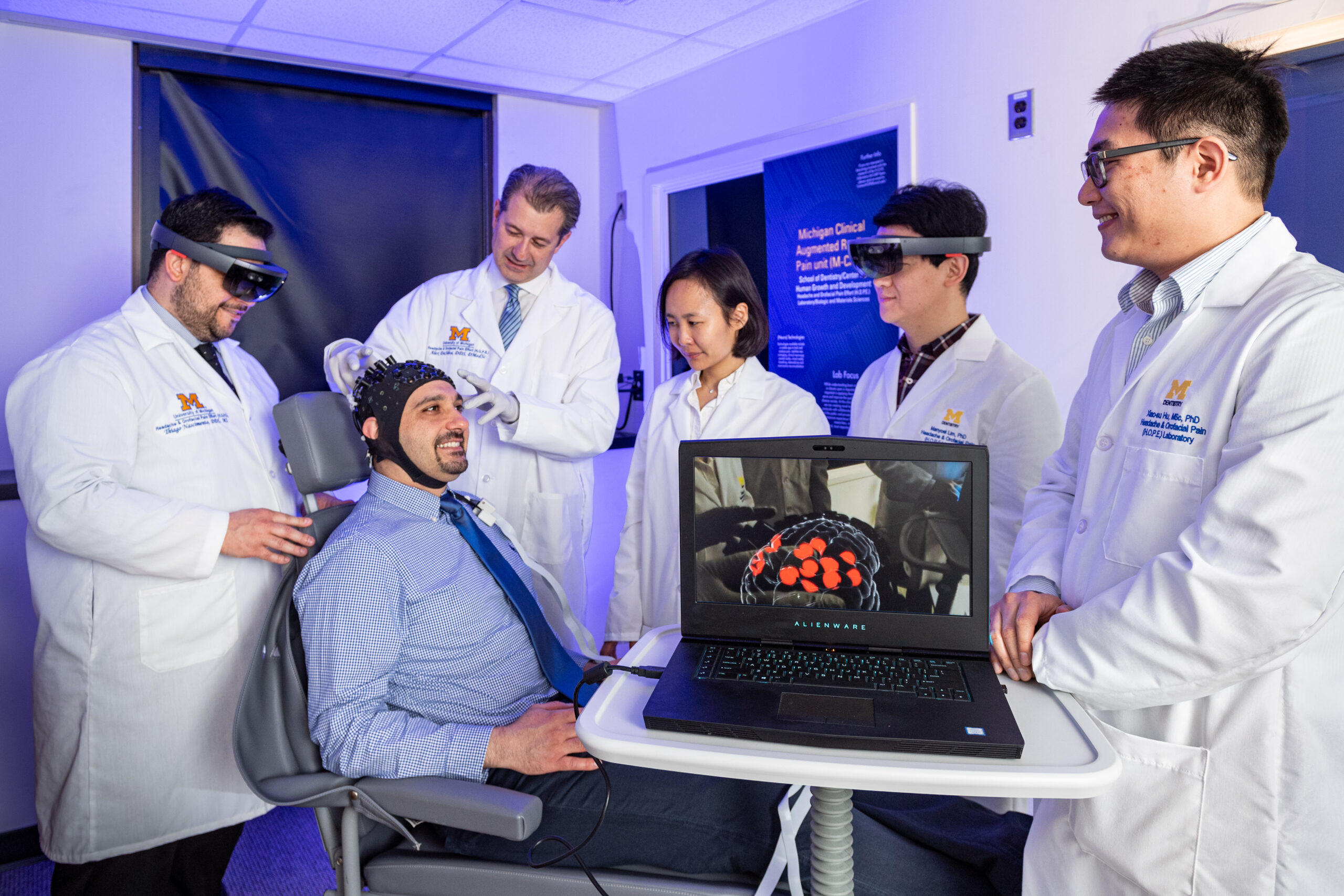 Hassan Jassar (seated) wears a sensor-outfitted cap that detects changes in blood flow and oxygenation, thus sensing brain activity. That information is transmitted to a computer and interpreted. Thiago Nascimento, left, views this brain activity in real-time while wearing augmented reality glasses, and the computer image shows that particular pain signature in the brain. From left to right, also pictured, Dr. Alex DaSilva, Dajung Kim, Manyoel Lim, Xiao-su Hu.