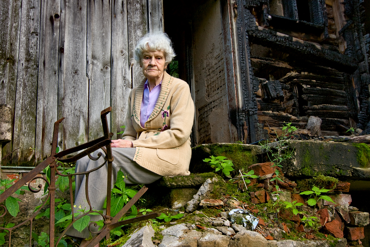Elderly woman sitting outside a ruined building. Image credit: iStock