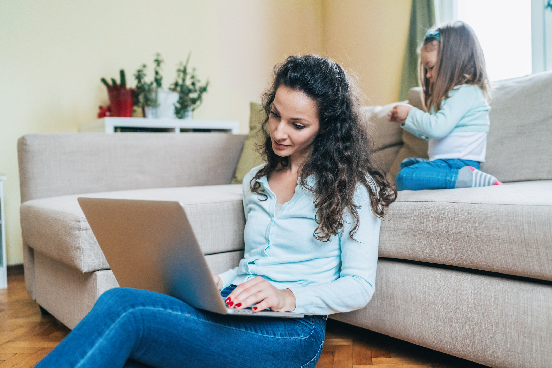 A mother uses a computer with a child in the background. Image credit: iStock