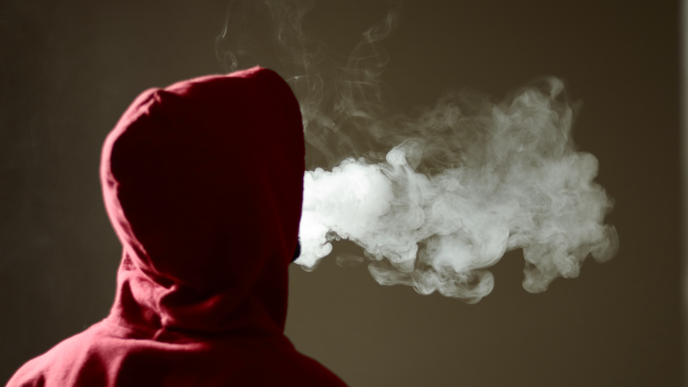 Young male in red hoodie vaping smoking, exhales thick vapor, isolated rear view. Image credit: property of Naveen Asaithambi