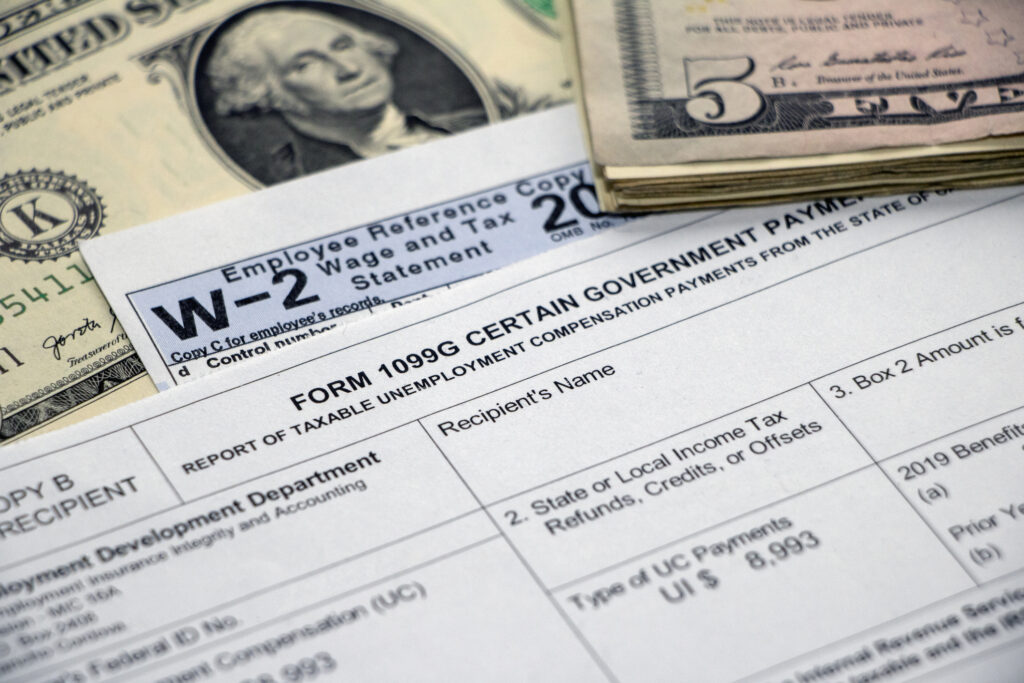 iStock Image: Closeup of a Form 1099G Certain Government Payments for unemployment benefits atop a W2 form and dollar bill, with stack of $5 bills.