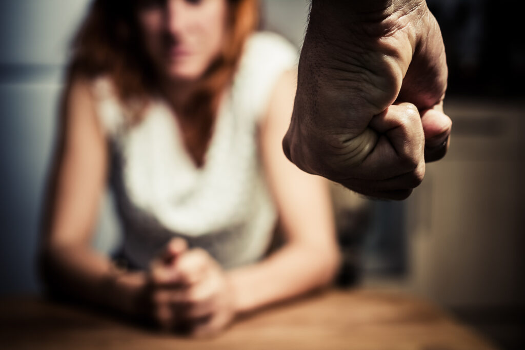 Young woman is sitting hunched at a table at home, the focus is on a man's fist in the foregound of the image. image credit: iStock