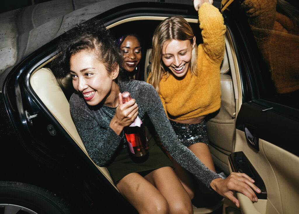 Women leaving backseat of cab with drinks in hand. Image credit: iStock