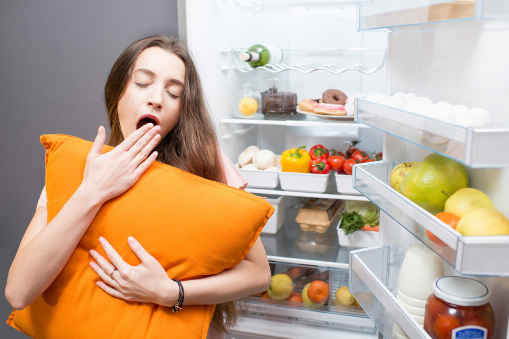 Woman with a pillow next to a refrigerator stock with fruits and veggies. Image credit: iStock