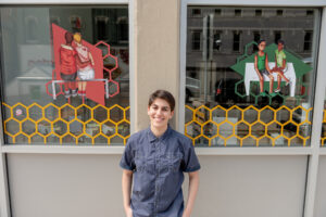 Shalin Berman, University of Michigan Stamps School of Art & Design student, poses with their art at Bløm Meadworks. Berman's work is part of "The art of queer health sciences," an exhibition on view at several downtown Ann Arbor businesses and at queeringart.com. Image credit: Eric Bronson/Michigan Photography.