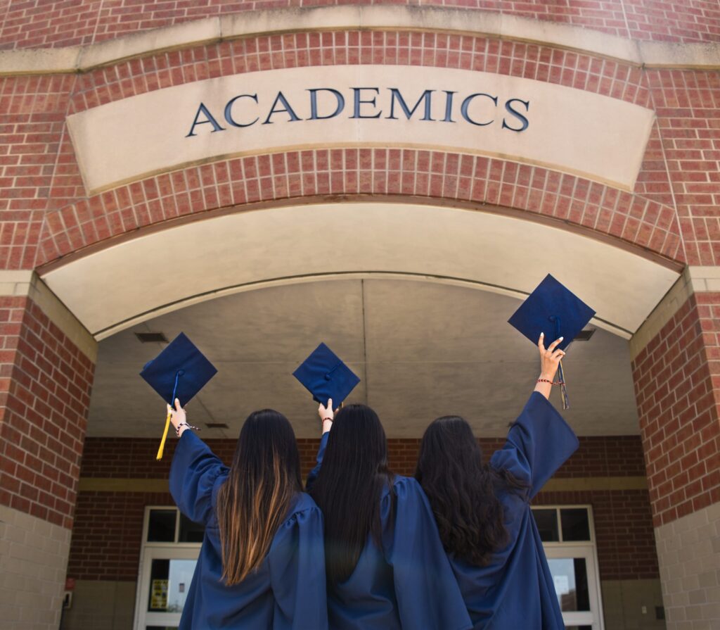 Female graduates raise hats at building sign. Image credit: Leon Wu, Unsplash