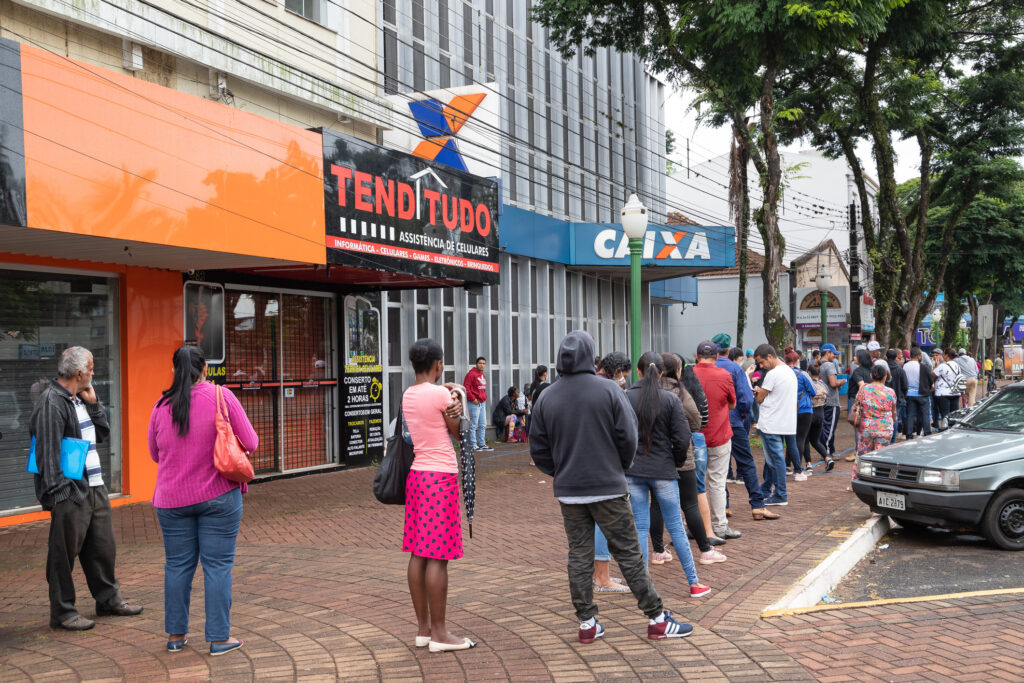 Mask-protected population awaits service outside a bank branch to receive emergency benefit from the Brazilian government during coronavirus pandemic. Image credit: iStock
