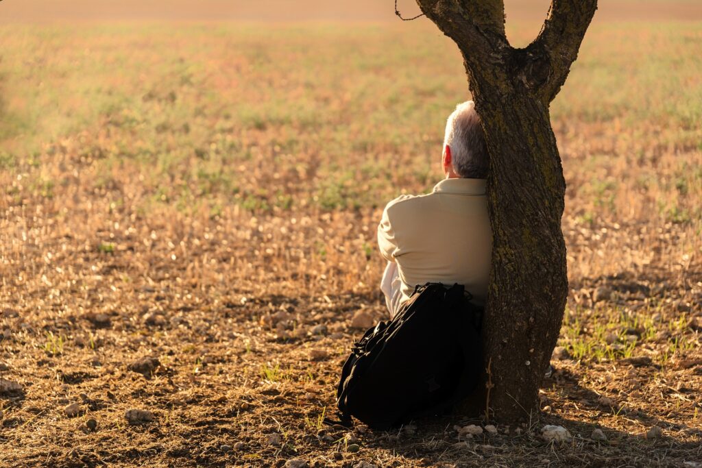 I man sits leaning against the base a a tree with a backpack beside him. Image credit: josealbafotos, Pixabay.com