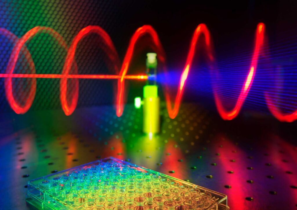 The illustration shows a microplate well in the foreground, while in the background a tested sample receives red laser light and releases twisted blue light. Image credit: Ventsislav Valev, Kylian Valev and Lukas Ohnoutek, University of Bath