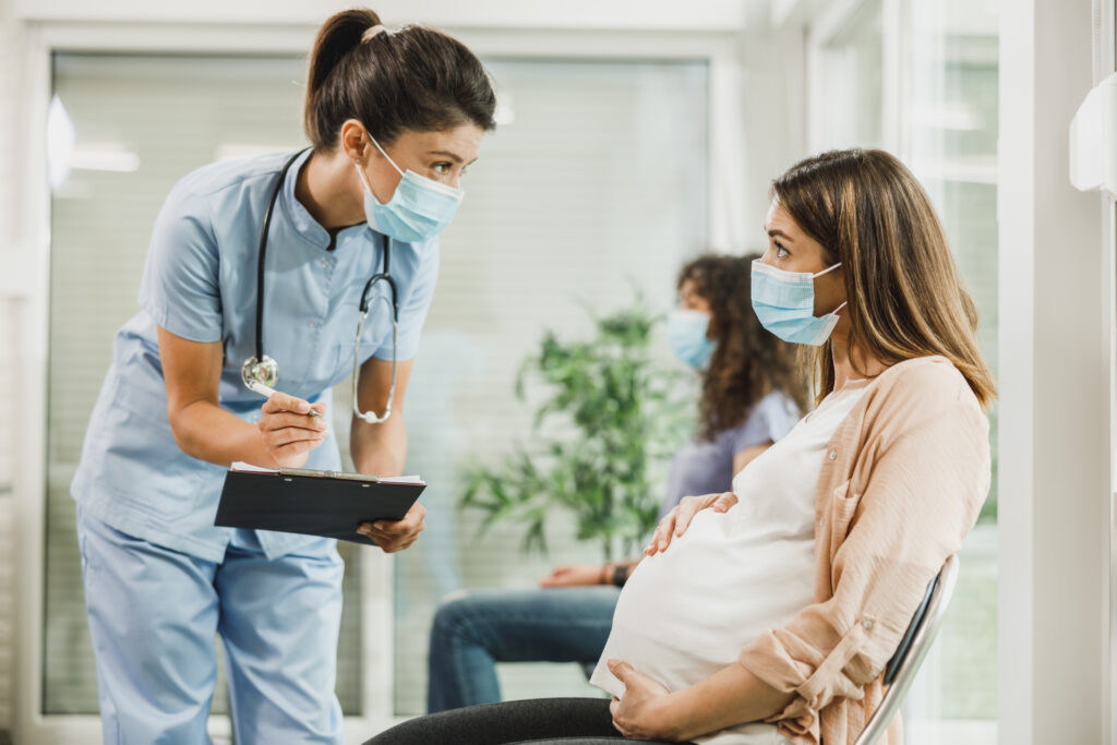 Pregnant Woman With Protective Mask Talking To Nurse At Waiting Room. Image credit: iStock