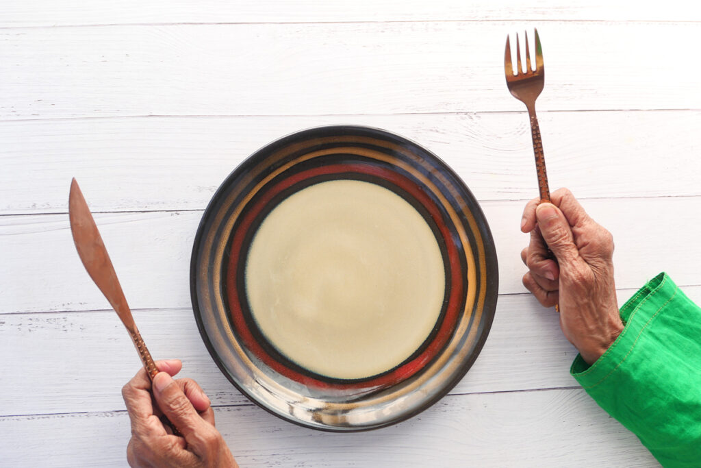 Elderly hands hold a fork and knife next to an empty plate. Image credit: iStock