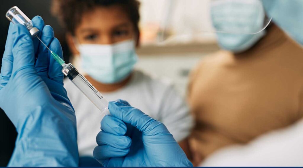Gloved hands drawing a vaccine with masked child and adult in the backgrounds. Image courtesy: DMACS