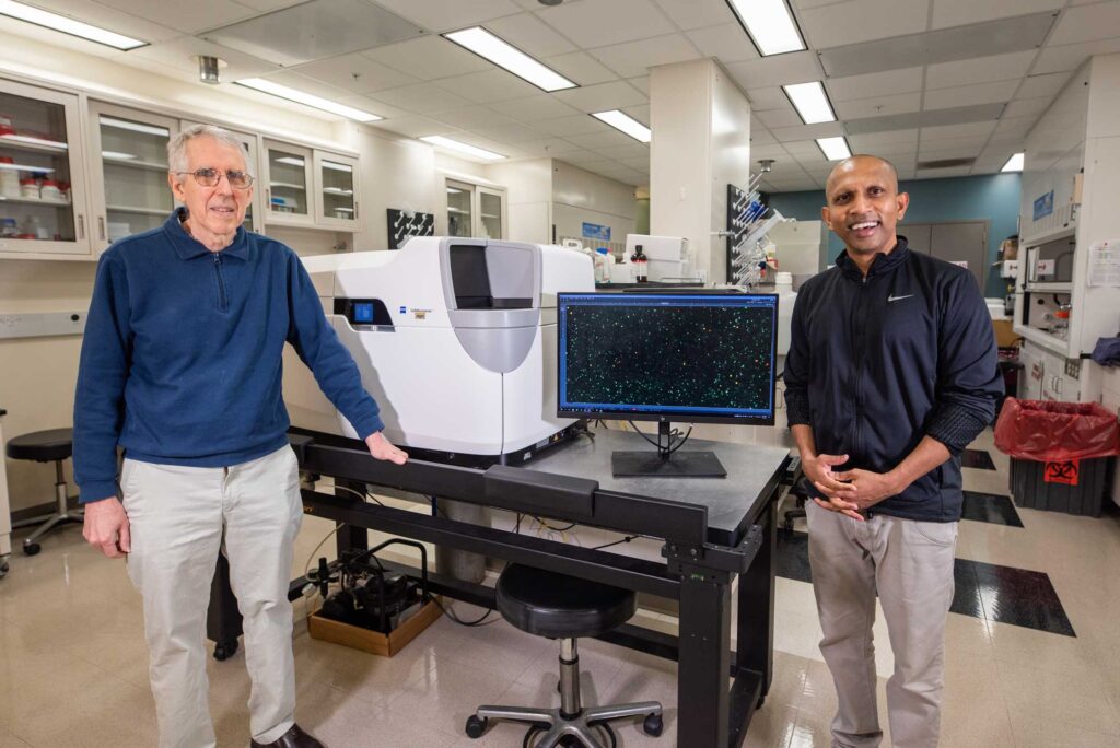 Hero and Rajapaske stand next to the microscope display showing an image of the multiplying B-cells. Image Credit: Silvia Cardarelli