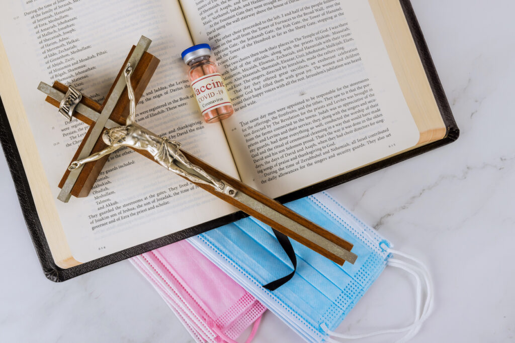 A crucifix and vaccine vial sitting on an open bible and masks. Image credit: iStock