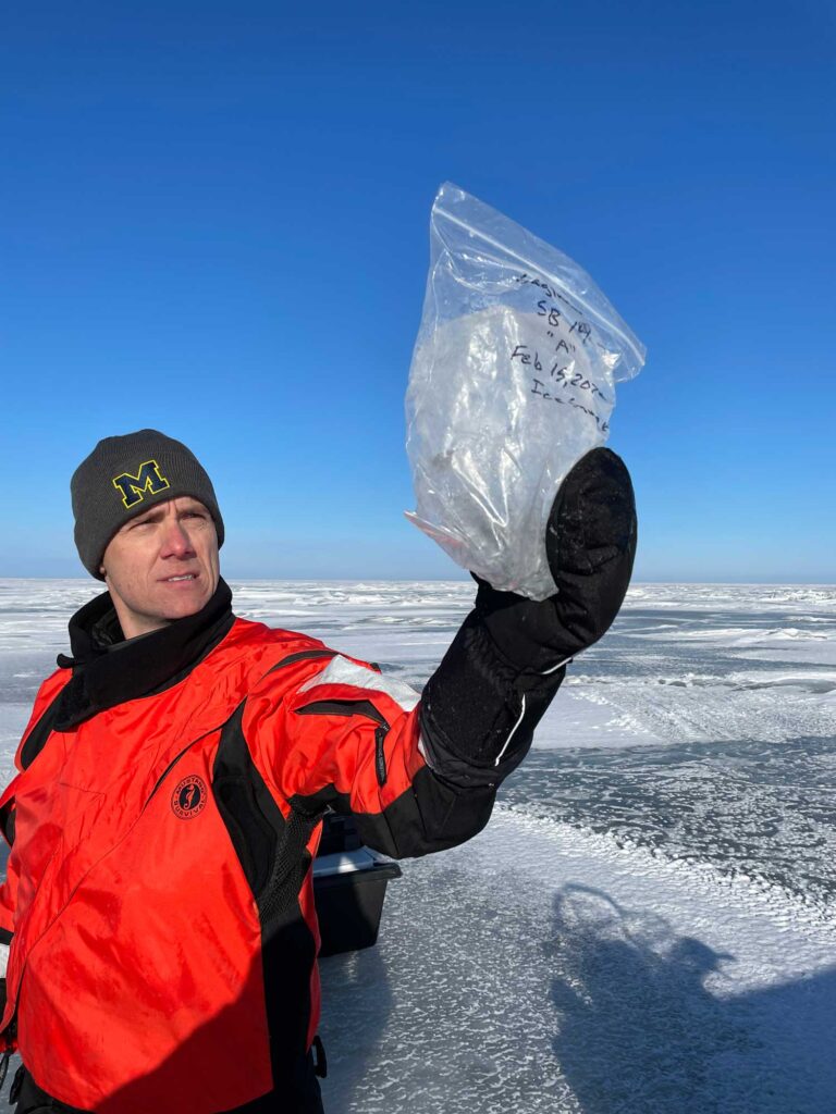 University of Michigan biogeochemist Casey Godwin raises a football-size chunk of crystal-clear Great Lakes ice toward the sky and studies it. The ice was plucked from the Saginaw Bay surface during a Feb. 15 trip to collect ice and water samples for the Winter Grab coordinated campaign. This sample was melted and used to measure dissolved substances in the ice. Image credit: Michigan News