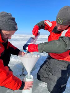 Andrew Camilleri, left, and Casey Godwin pour lake water through a funnel and into storage bottles. The white PVC tube, called a Van Dorn Sampler, was used to collect water from known depths. Image credit: Michigan News