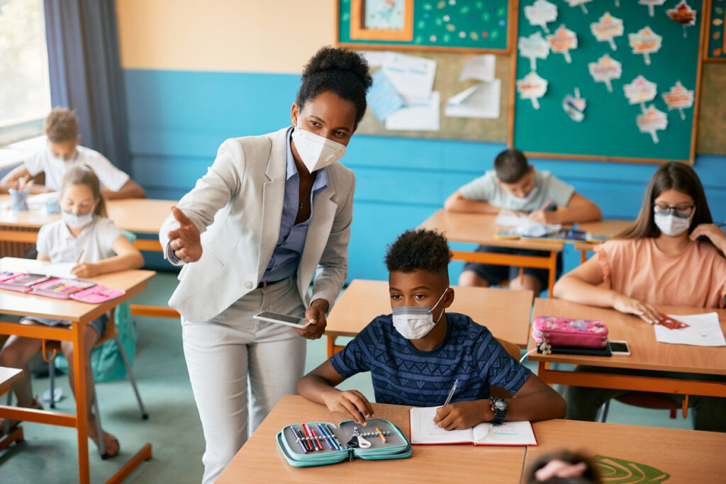 Elementary school teacher and her students wearing protective face mask in the classroom. Image credit: iStock