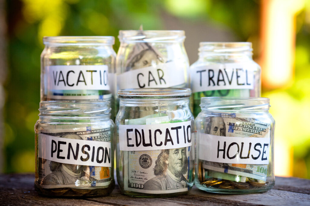 A lot coins in glass money jar on the wood table. Saving for house, health, education, car  and travel concept. Image credit: iStock