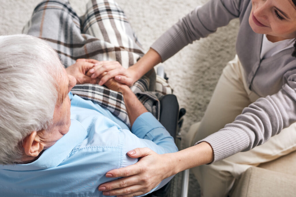 Young supportive female caregiver sitting by senior man in wheelchair and keeping her hand on his shoulder while comforting him. Image credit: iStock
