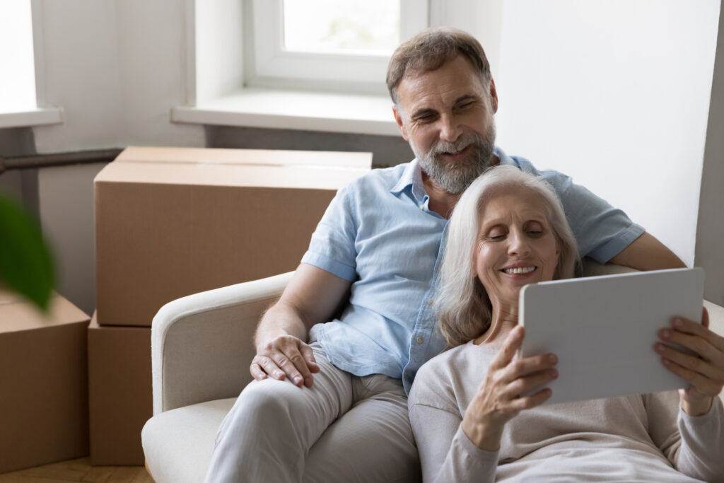 Older couple relax on sofa on move day near boxes with stuff, use tablet buy goods on internet, make order via e-service app, make purchase online, search home renovation new ideas, e-commerce concept. Image credit: fizkes