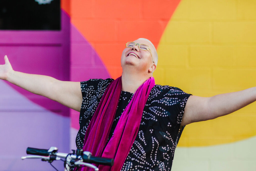 Petra Kuppers, the Anita Gonzalez Collegiate Professor of Performance Studies and Disability Culture in LSA, smiles up to the sky, arms outstretched, embracing the world. Her mobility scooter’s handlebar is visible at the bottom of the image. (Photo by Tamara Wade)