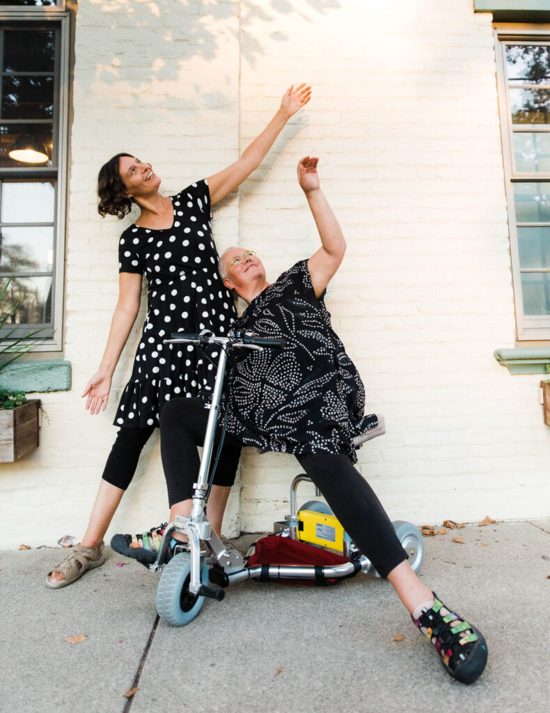 Petra Kuppers, with yellow glasses, and her wife, Stephanie Heit, lean back against a cream-colored house wall, and reach upward with their left arms, smiling, in contact and community play. (Photo by Tamara Wade)