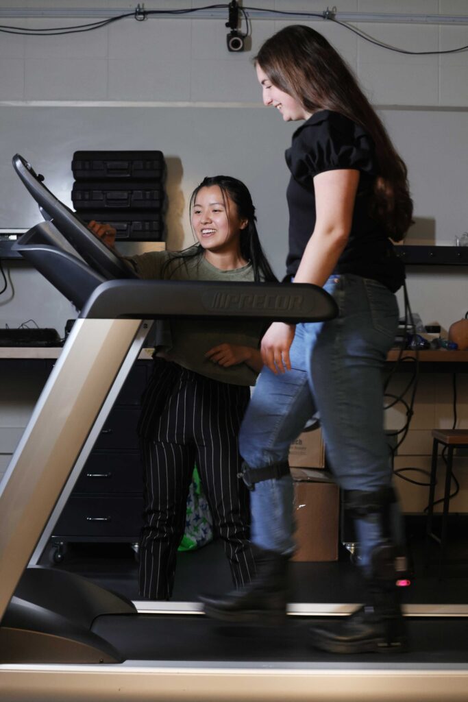 Man I (Maggie) Wu, a PhD student in robotics, left, talks with Jacqueline Hannan, a PhD student in industrial and operations engineering, as they test out an ankle exoskeleton for another project in Stirling's lab. Photo credit: Brenda Ahearn, University of Michigan Engineering