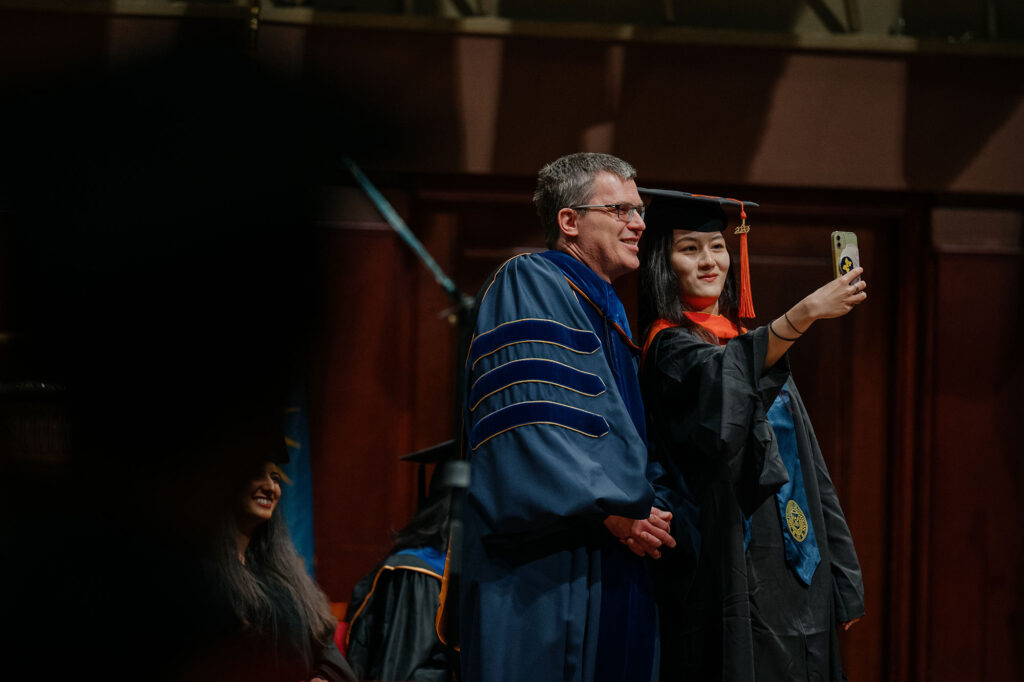 Photo of a master's degree graduate taking a selfie with Rackham Dean Michael Solomon
