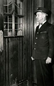 Black & white photo of doorman of the Michigan Union, George Johnson. He is standing at the inside of the front door of the Michigan Union. (Image courtesy of U-M's Bentley Historical Library)
