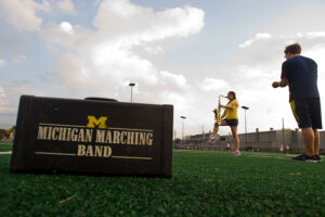 A horn player is silhouetted against the evening sky with a band case in the foreground that reads Michigan Marching Band with a Block M.