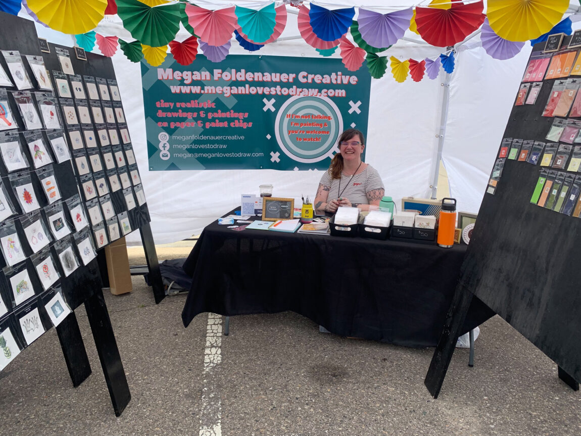 Megan Foldenauer, multimedia specialist for Michigan Medicine Neurosurgery, sits among many of the works of art in her booth at the Ann Arbor Art Fair. (Photo courtesy of Megan Foldenauer)
