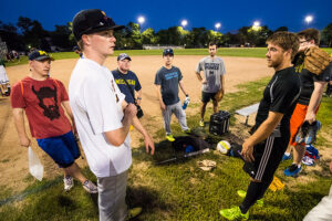 An intramural baseball team talks strategy on the sidelines. Young men in a semi-circle dressed in mismatching t-shirts.