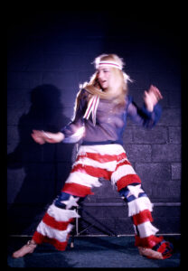 Pat Oleszko as the Hippie Strippy. She's dressed in a see-through top with American flag fringe pants.