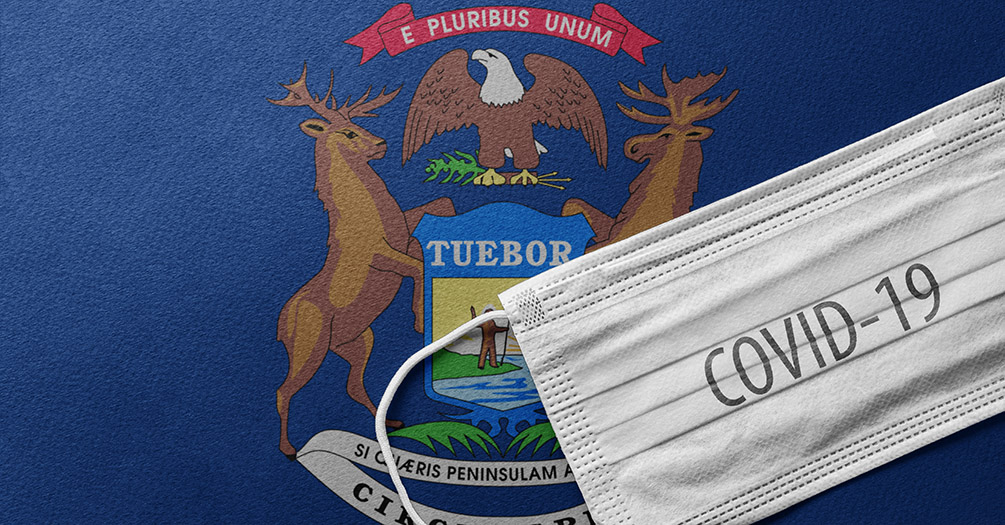 A surgical mask with the words COVID-19 sits on top of at Michigan flag. Image credit: iStock