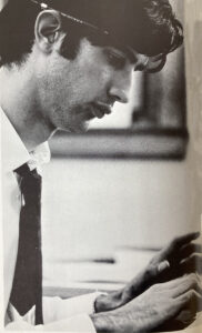 Roger Rapoport, BA '68, at his desk at the Michigan Daily. He's wearing a white button down shirt with a tie. And has a pencil tucked behind his ear.