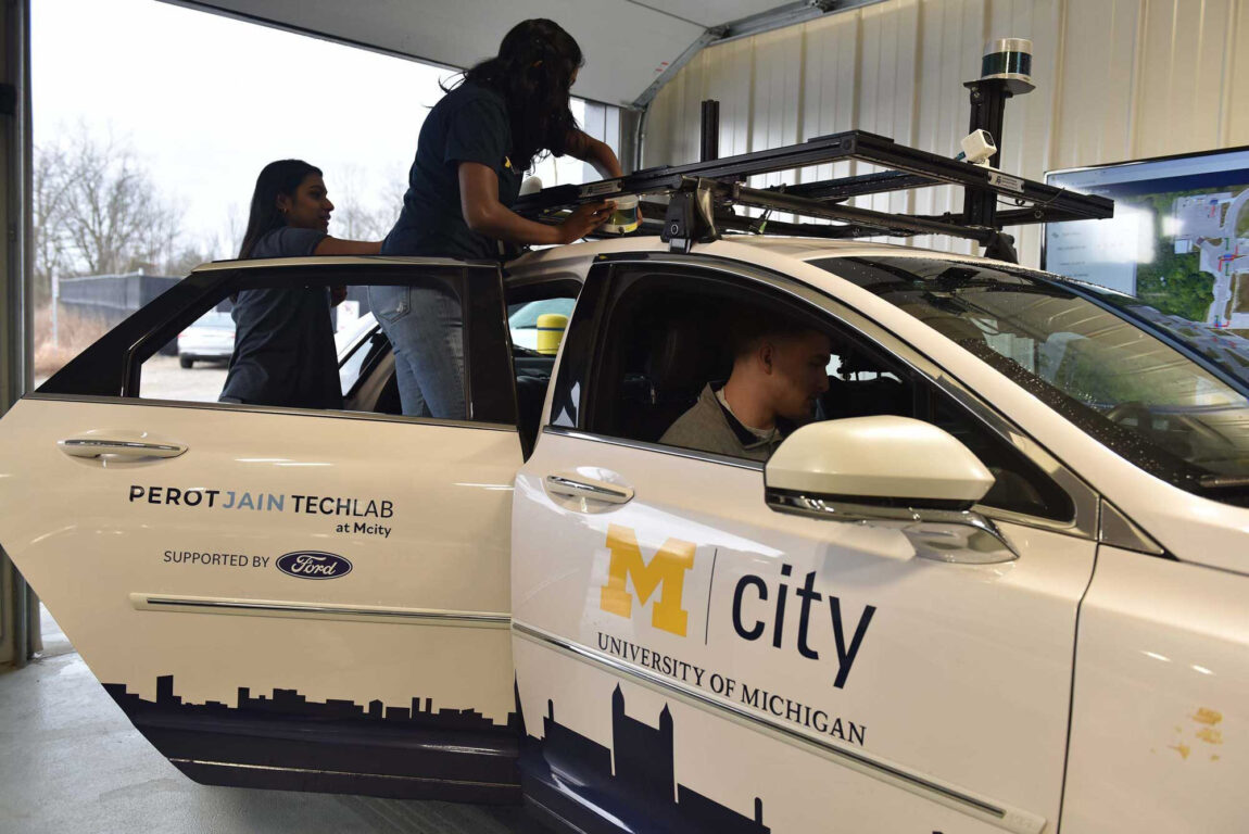 A photo showing Neha Tiwari and Swetha Subbiah mounting a LiDAR system on an Mcity vehicle while Broderick Riopelle ensures that the sensor is being read by the vehicle.