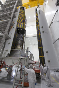 The Parker Solar Probe appears as a gunmetal gray cylinder attached to a cone-shaped base. The probe is nested inside half of a tall, rocket-shaped case. The outside of the case is white and smooth while the inside is lined with many black panels with yellow borders. Together, the panels create a checkerboard pattern. The probe is resting on a raised platform, and the other half of the casing faces the opposite side of the probe. Two scientists work around the probe on the raised platform while three others work below.