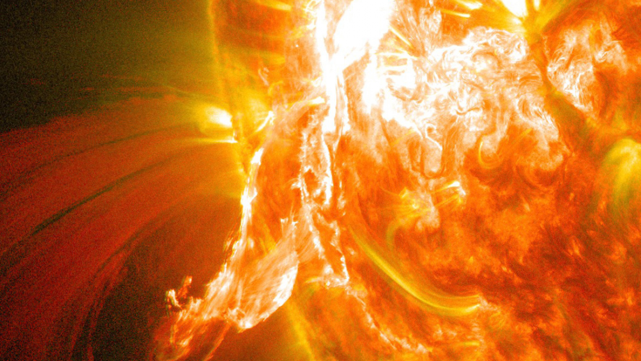 The sun is viewed from space and appears as a fiery red and orange sphere covering more than half of the image. A red and orange arc of plasma, which resembles fire, protrudes from the sun’s surface towards the bottom of the frame. The plasma extends from a very bright white section of the sun’s surface. Image credit: NASA/Solar Dynamics Observatory